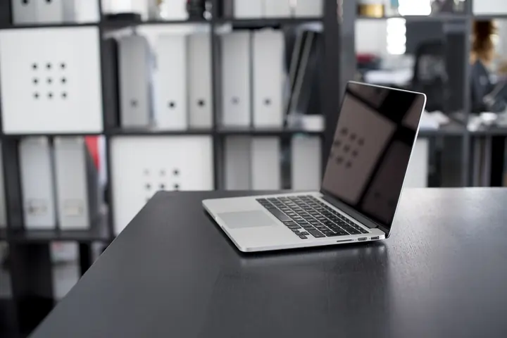 A laptop open on a tidy office desk in front of shelves of box files.